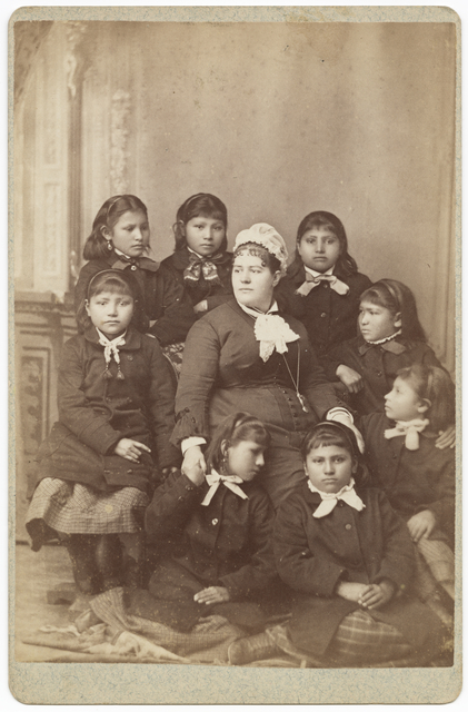 Sepia photo of seated teacher surrounded by 8 Native American girls